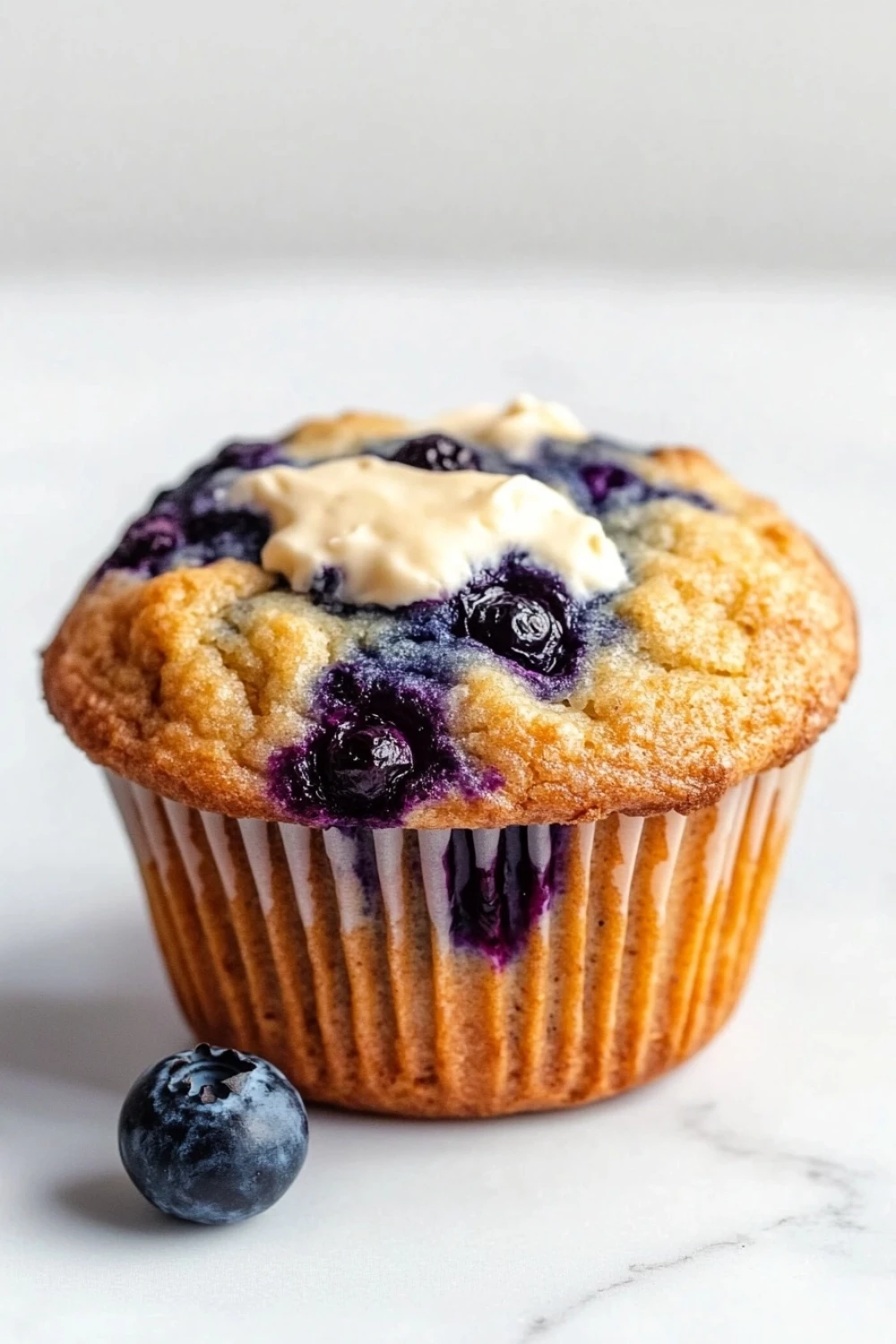 Blueberry muffin with cream cheese - the image is of a freshly baked blueberry muffin on a white marble countertop. the muffin has a golden brown crust and is topped with fresh blueberries and a dollop of white cream cheese frosting. there are two whole blueberries on the countertop next to the muffin. the background is blurred, making the blueberries the focal point of the image.