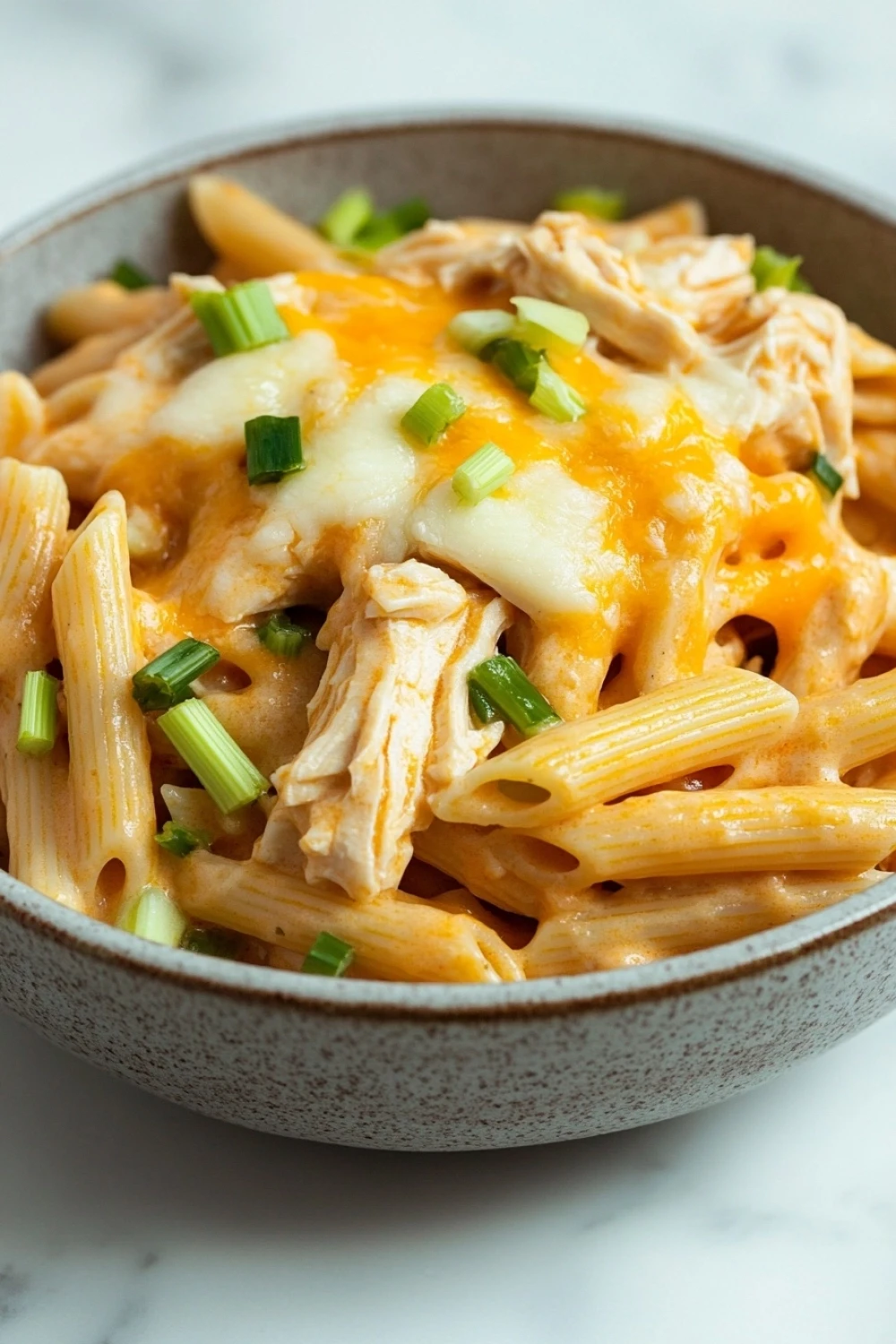 Buffalo chicken pasta freezer - the image is a close-up of a bowl of pasta dish. the pasta appears to be penne, which is a type of penne pasta, and is covered in a creamy orange sauce with chunks of chicken and melted cheese on top. the sauce is drizzled over the top of the pasta, creating a smooth and creamy texture. there are also some chopped green onions scattered throughout the dish, adding a pop of color. the bowl is made of ceramic and is sitting on a white marble countertop.