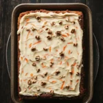 Carrot cake in 9 x 13 pan - the image shows a rectangular baking dish with a layer of carrot cake in it. the cake appears to be freshly baked and has a golden brown crust on top. the top of the cake is covered in a creamy white frosting with chopped nuts scattered throughout. the frosting is drizzled over the cake, giving it a smooth and shiny appearance. the dish is sitting on a dark wooden table with a black plate underneath.