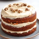 Carrot cake no eggs - the image is of a round cake on a white marble countertop. the cake appears to be a carrot cake with a layer of white frosting on top. the frosting is drizzled with chopped pecans, giving the cake a crunchy texture. the top of the cake is covered in a light dusting of powdered sugar. in the background, there is another cake on another plate, suggesting that the cake has been freshly baked.