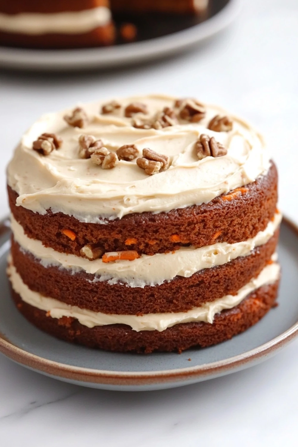 Carrot cake no eggs - the image is of a round cake on a white marble countertop. the cake appears to be a carrot cake with a layer of white frosting on top. the frosting is drizzled with chopped pecans, giving the cake a crunchy texture. the top of the cake is covered in a light dusting of powdered sugar. in the background, there is another cake on another plate, suggesting that the cake has been freshly baked.