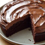 Chocolate cake for kids - the image is of a chocolate cake on a white plate. the cake has a round shape and is covered in a thick layer of chocolate frosting. the frosting is swirled in a zigzag pattern and appears to be smooth and glossy. a slice of the cake is cut out of the plate, revealing the moist, chocolate cake inside. the plate is sitting on a wooden table.