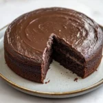 Chocolate cake old fashioned - the image shows a round chocolate cake on a white marble plate. the cake has a thick layer of chocolate frosting on top and a slice taken out of it. the frosting is smooth and shiny, and it appears to be freshly baked. the plate is round and has a gold rim. the background is blurred, but it seems to be a kitchen countertop.