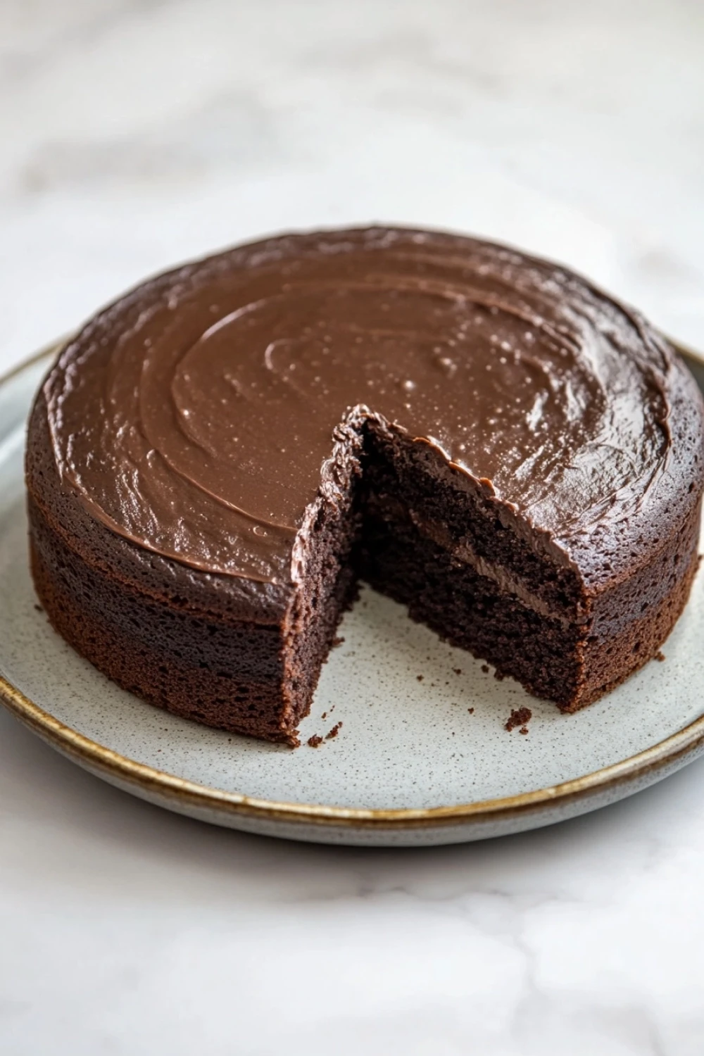 Chocolate cake old fashioned - the image shows a round chocolate cake on a white marble plate. the cake has a thick layer of chocolate frosting on top and a slice taken out of it. the frosting is smooth and shiny, and it appears to be freshly baked. the plate is round and has a gold rim. the background is blurred, but it seems to be a kitchen countertop.
