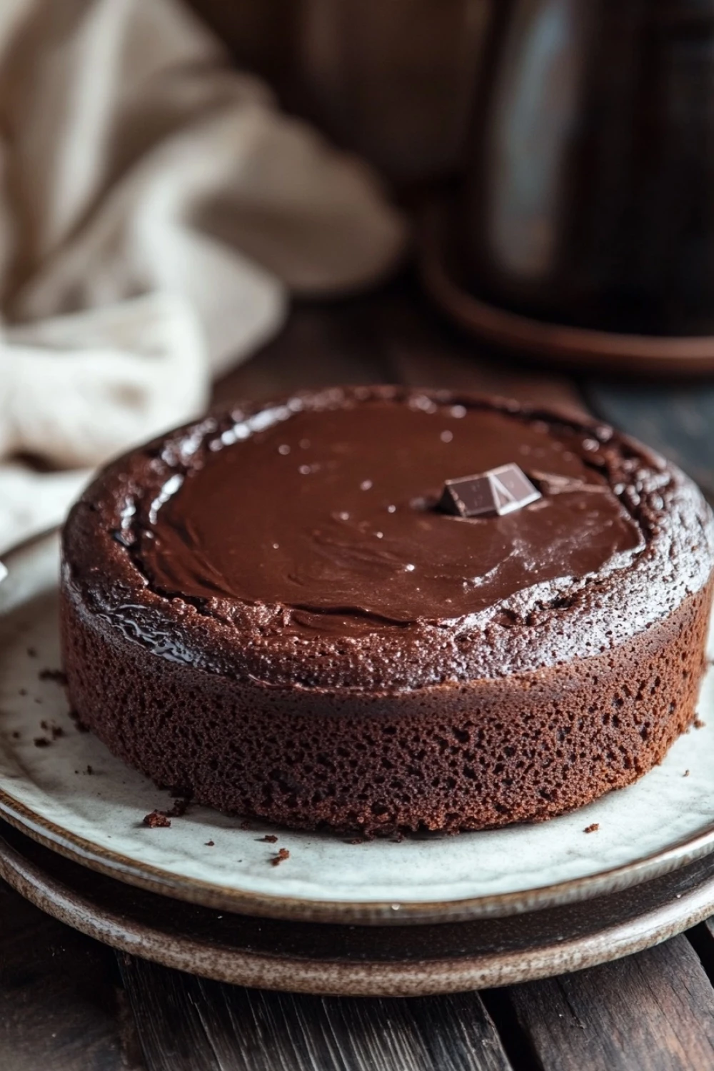 Chocolate cake photography - the image shows a round chocolate cake on a white plate. the cake is covered in a thick layer of chocolate frosting and has a small chocolate bar on top. the plate is placed on a wooden table with a white cloth napkin and a teapot in the background. the overall mood of the image is rustic and cozy.