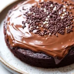 Chocolate cake toppings - the image is of a round chocolate cake on a white plate. the cake is covered in a thick layer of chocolate ganache, which is drizzled over the top of the cake. there are small white sprinkles scattered around the cake, adding a pop of color to the dish. the plate has a speckled pattern around the edges. the background is a plain white surface.