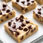 Chocolate chip cookie dough bars - the image shows a tray of freshly baked chocolate chip cookie bars. the bars are golden brown in color and have a crumbly texture. they are arranged in a single layer on the tray, with some of the bars overlapping each other. the chocolate chips are scattered on top of the cookie bars, adding a pop of color and texture. the tray is lined with parchment paper, and the background is a light blue color.