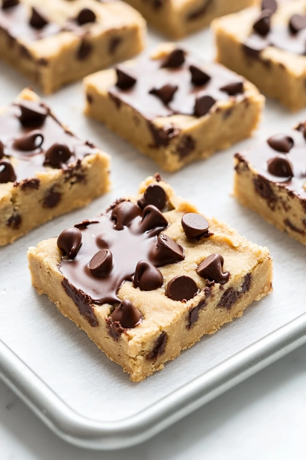 Chocolate chip cookie dough bars - the image shows a tray of freshly baked chocolate chip cookie bars. the bars are golden brown in color and have a crumbly texture. they are arranged in a single layer on the tray, with some of the bars overlapping each other. the chocolate chips are scattered on top of the cookie bars, adding a pop of color and texture. the tray is lined with parchment paper, and the background is a light blue color.