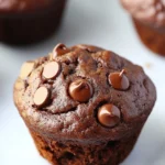 Chocolate muffin no egg - the image shows a close-up of a freshly baked chocolate muffin on a white parchment paper. the muffin is round and has a golden brown color. it is topped with chocolate chips and drizzled with chocolate sauce. in the background, there are more muffins visible, but they are slightly blurred. a knife is resting on the parchment paper next to the muffin.