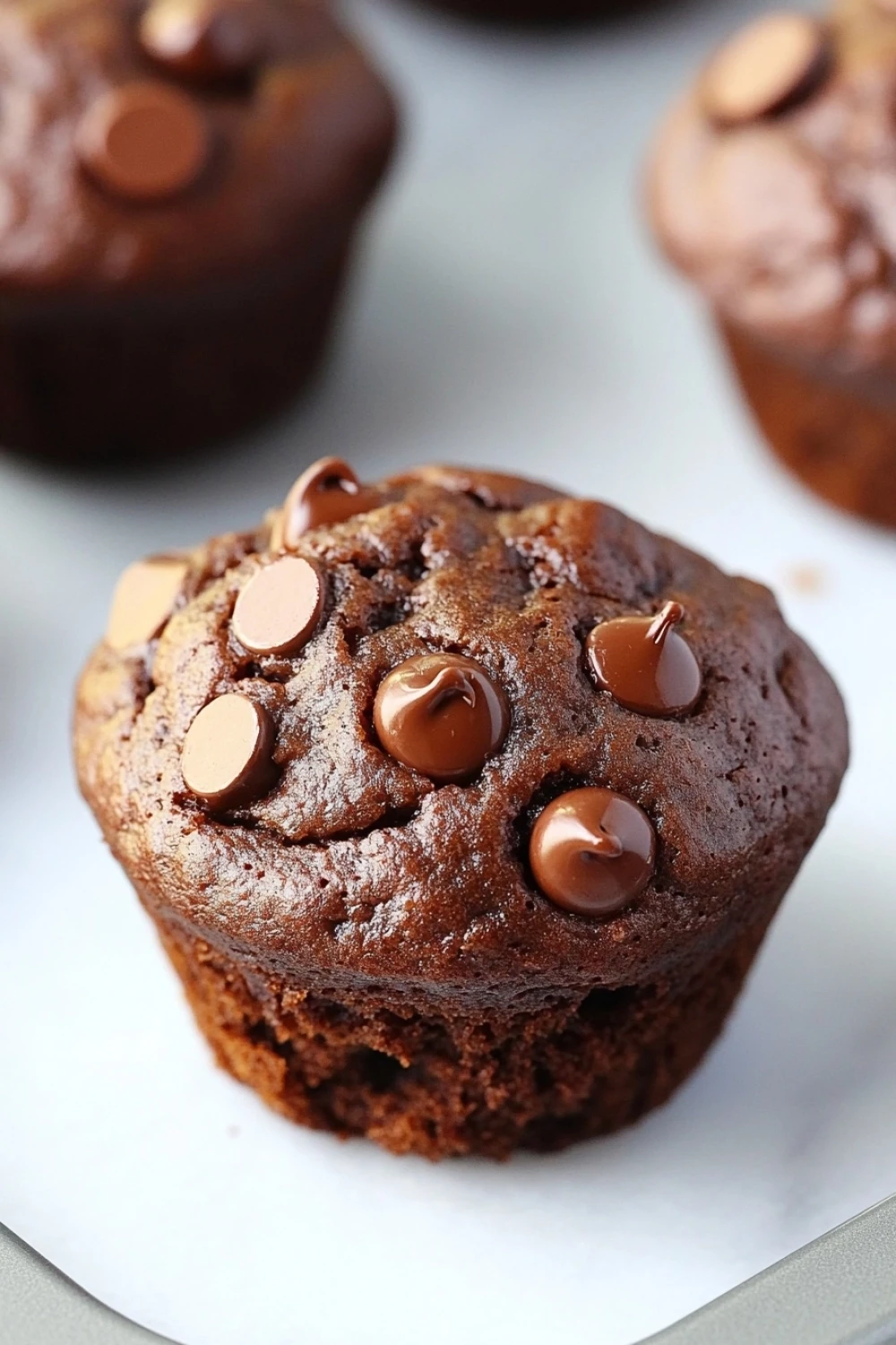 Chocolate muffin no egg - the image shows a close-up of a freshly baked chocolate muffin on a white parchment paper. the muffin is round and has a golden brown color. it is topped with chocolate chips and drizzled with chocolate sauce. in the background, there are more muffins visible, but they are slightly blurred. a knife is resting on the parchment paper next to the muffin.