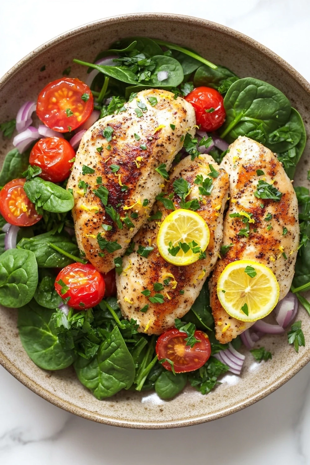 Eating well weeknight lemon chicken skillet dinner - the image is a close-up of a plate of food. the plate is made of a light-colored ceramic bowl and is placed on a white marble countertop. on top of the salad, there are two pieces of grilled chicken breasts that are golden brown and appear to be seasoned with herbs and spices. the chicken is garnished with sliced lemons and red onions. the salad is made up of fresh spinach leaves, cherry tomatoes, and red onion slices. the overall color scheme of the dish is vibrant and appetizing.