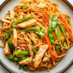 Freezer chicken lo mein - the image is a close-up of a plate of noodles with chicken and vegetables. the noodles are orange in color and appear to be stir-fried. the chicken is cut into small pieces and is arranged in a circular pattern on top of the noodles. the vegetables include sliced carrots, green bell peppers, and snow peas. the dish is garnished with sesame seeds. the plate is sitting on a white marble countertop.