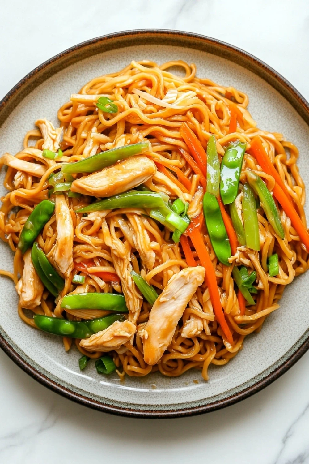 Freezer chicken lo mein - the image is a close-up of a plate of noodles with chicken and vegetables. the noodles are orange in color and appear to be stir-fried. the chicken is cut into small pieces and is arranged in a circular pattern on top of the noodles. the vegetables include sliced carrots, green bell peppers, and snow peas. the dish is garnished with sesame seeds. the plate is sitting on a white marble countertop.