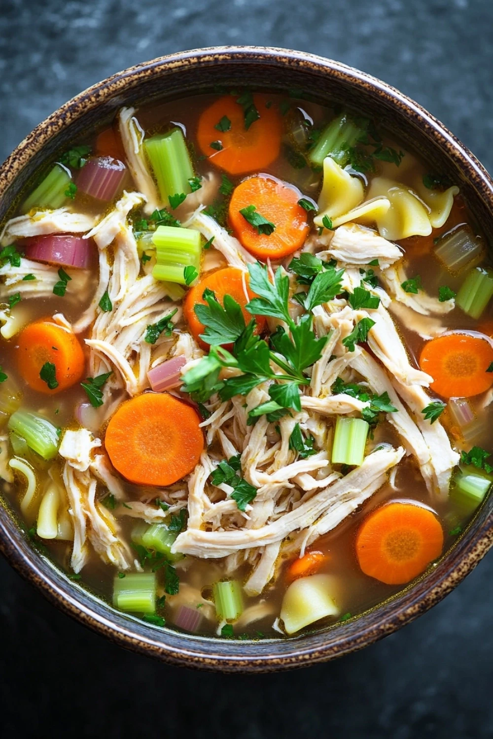 Freezer chicken soup - the image is a close-up of a bowl of soup. the soup appears to be made with chicken, carrots, celery, and other vegetables. the bowl is made of ceramic and has a dark brown color. the broth is thick and creamy, and the vegetables are bright orange and green. there are also chunks of chicken and celery scattered throughout the soup, and a sprig of parsley is visible on top. the background is a dark grey countertop.