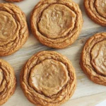 Gingerbread cookie chewy - the image shows a wooden cutting board with twelve round cookies arranged in a circular pattern. the cookies appear to be freshly baked and have a golden brown color. they are arranged in two rows of three, with the top row slightly overlapping the bottom row. each cookie has a swirl pattern on top, with some of the cookies overlapping each other. the background is a white wooden surface, and the focus is on the cookies.