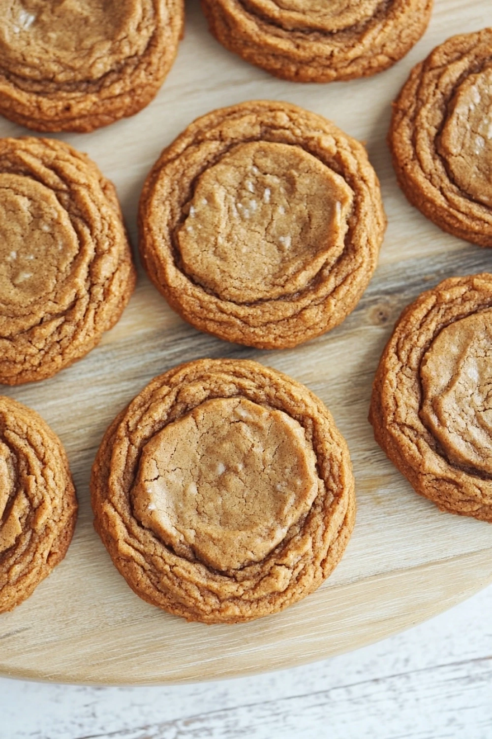 Gingerbread cookie chewy - the image shows a wooden cutting board with twelve round cookies arranged in a circular pattern. the cookies appear to be freshly baked and have a golden brown color. they are arranged in two rows of three, with the top row slightly overlapping the bottom row. each cookie has a swirl pattern on top, with some of the cookies overlapping each other. the background is a white wooden surface, and the focus is on the cookies.