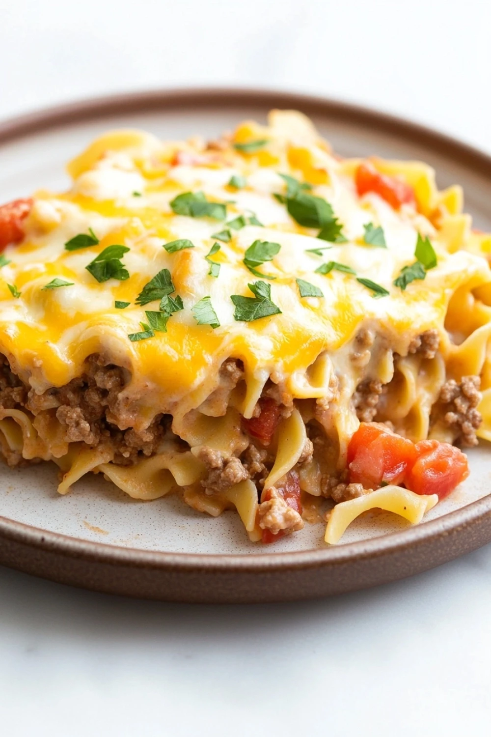 Ground beef casserole with noodles - the image shows a plate of lasagna on a white tablecloth. the lasagna is made with fettuccine noodles, ground beef, and tomato sauce. the top of the lasagna has melted cheese and garnished with fresh parsley. the plate is brown and round, and the dish looks freshly cooked and ready to eat.