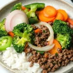 Ground beef dinner for one - the image is a close-up of a plate of food. the plate is white with a light brown rim and is placed on a white marble countertop. the food appears to be a stir-fry dish with ground beef, carrots, broccoli, red onions, and green bell peppers. the beef is cooked to a medium-rare and the vegetables are arranged in a colorful and appetizing manner. the rice is white and fluffy, and the plate is garnished with a sprinkle of sesame seeds.