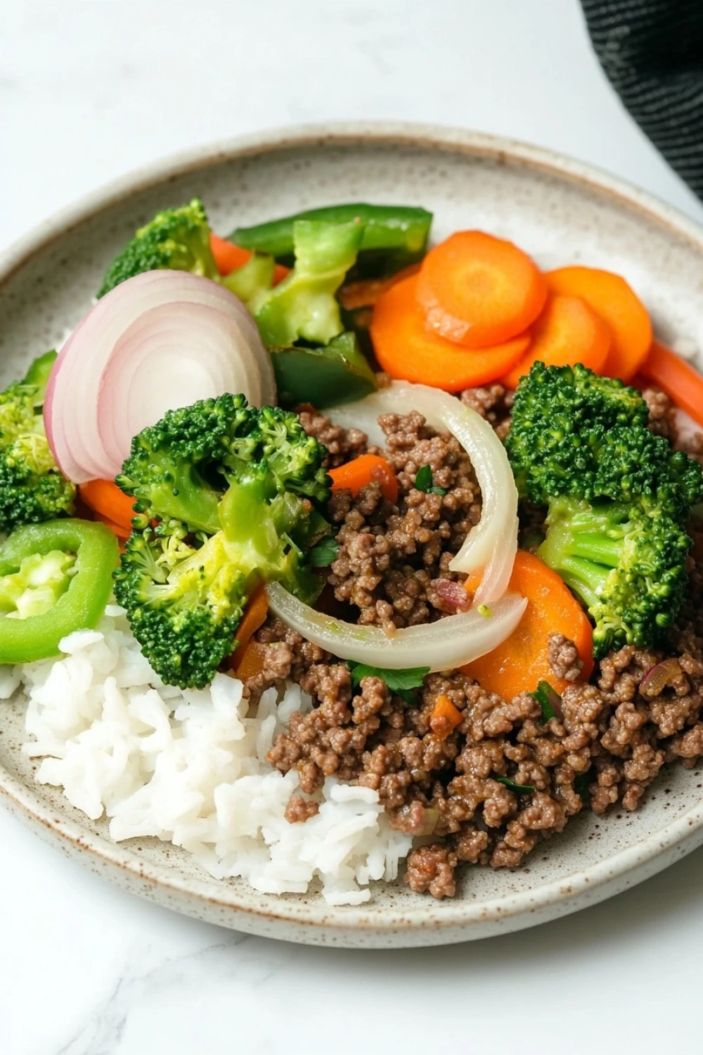 Ground beef dinner for one - the image is a close-up of a plate of food. the plate is white with a light brown rim and is placed on a white marble countertop. the food appears to be a stir-fry dish with ground beef, carrots, broccoli, red onions, and green bell peppers. the beef is cooked to a medium-rare and the vegetables are arranged in a colorful and appetizing manner. the rice is white and fluffy, and the plate is garnished with a sprinkle of sesame seeds.