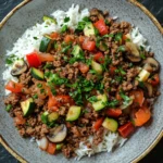 Ground beef dinner no dairy - the image is a close-up of a bowl of food. the bowl is made of ceramic and has a brown rim. the food in the bowl appears to be a dish made with ground beef, sliced vegetables, and mushrooms. the vegetables include zucchini, carrots, and bell peppers. the rice is white and fluffy, and the dish is garnished with chopped parsley. the background is a dark grey textured surface.