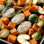 Harvest chicken sausage sheet pan - the image shows a tray of roasted vegetables on a black table. the vegetables are arranged in a circular pattern on the tray, with some overlapping each other. on top of the vegetables, there are roasted sweet potatoes, brussels sprouts, apples, and rosemary sprigs. in the center of the tray is a piece of cooked chicken, which appears to be seasoned with herbs and spices. the chicken is golden brown and looks crispy on the outside. the brussel sprouts are cut into small cubes and are arranged around the chicken, while the apples are sliced into thin slices. the rosemary is a light green color and is placed on top of each other, adding a pop of color to the dish.