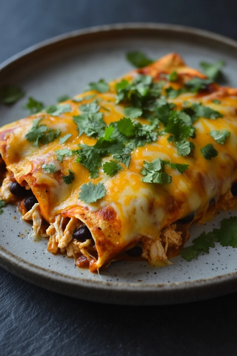 High protein chicken enchiladas - the image shows a plate of enchiladas on a dark grey plate. the enchillas are covered in melted cheese and garnished with fresh cilantro. the cheese is golden brown and appears to be melted and bubbly. there are also chunks of black beans scattered throughout the enchiles. the plate is sitting on a wooden table with a dark background.