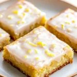 Lemon cake mix bars - the image shows four square-shaped cake bars on a white plate. the cake bars are golden brown in color and have a crumbly texture. they are topped with a generous layer of white frosting that is drizzled with lemon zest. the frosting is spread evenly over the top of the cake bars, creating a smooth and shiny surface. the plate is sitting on a wooden table with a light-colored background.
