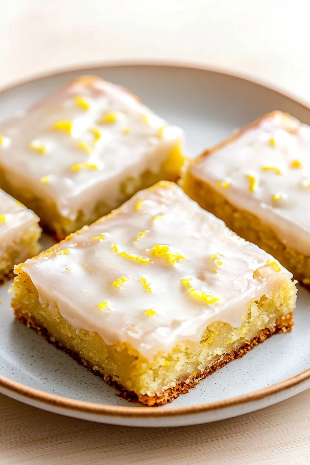 Lemon cake mix bars - the image shows four square-shaped cake bars on a white plate. the cake bars are golden brown in color and have a crumbly texture. they are topped with a generous layer of white frosting that is drizzled with lemon zest. the frosting is spread evenly over the top of the cake bars, creating a smooth and shiny surface. the plate is sitting on a wooden table with a light-colored background.