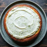 Lemon zucchini cake with cream cheese frosting - the image is of a round cake on a blue plate. the cake appears to be freshly baked and has a golden brown crust. on top of the cake, there is a generous layer of white frosting that is spread evenly across the cake. the frosting has small green herbs sprinkled on top, adding a pop of color to the dish. the plate is placed on a wooden table with a rustic texture.