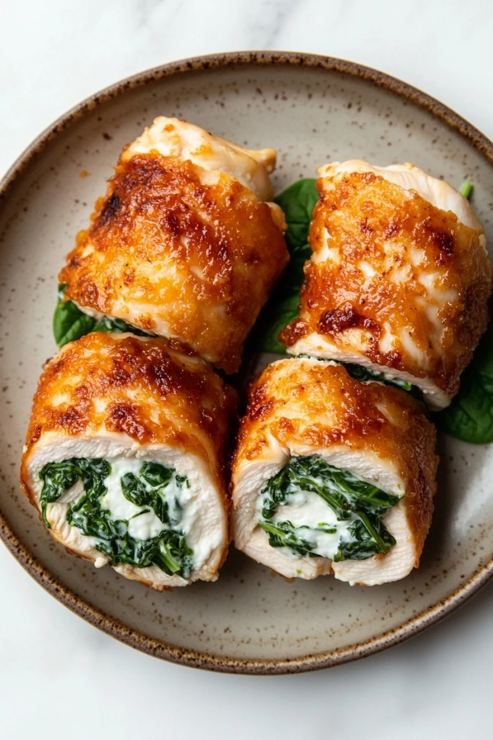 Low calorie chicken roll ups - the image shows a plate with four pieces of stuffed chicken breasts. the chicken breasts are golden brown and appear to be crispy on the outside. they are filled with a creamy white sauce and garnished with fresh spinach leaves. the plate is round and has a light brown rim. the background is a white marble countertop.
