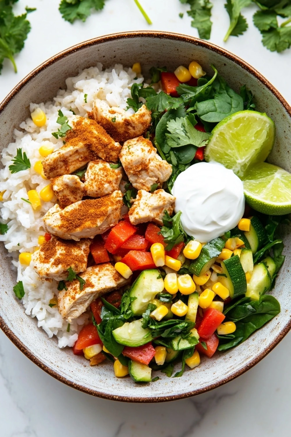 Make ahead chicken rice bowls - the image is a close-up of a bowl of food. the bowl is made of ceramic and is placed on a white marble countertop. the food in the bowl appears to be a salad with rice, chicken, corn, and vegetables. the chicken is cooked to a golden brown color and is garnished with fresh cilantro and lime wedges. there is also a dollop of sour cream on top of the salad. the salad is made up of sliced cucumbers, red bell peppers, and yellow corn kernels. the overall color scheme of the dish is vibrant and colorful.
