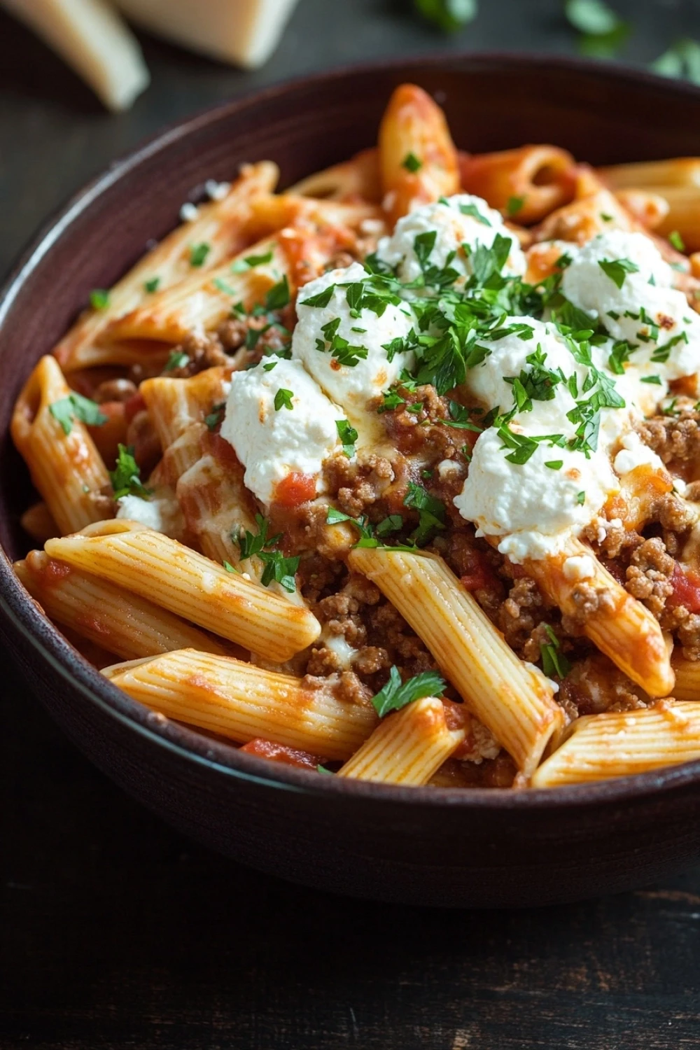 Make ahead dinners pasta - the image shows a bowl of penne pasta dish. the pasta is cooked al dente and is topped with a generous amount of ground beef and a dollop of sour cream. the dish is garnished with fresh parsley and there are a few pieces of parmesan cheese scattered around the bowl. the bowl is made of dark brown ceramic and is sitting on a dark wooden table. the background is blurred, but it appears to be a kitchen countertop.