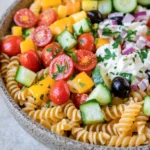 Make ahead pasta salad - the image is a close-up of a bowl of pasta salad. the bowl is made of ceramic and has a light brown color. the pasta is fusilli, which is a type of spiral-shaped pasta, and it is mixed with various vegetables such as sliced cucumbers, red onions, cherry tomatoes, and black olives. the vegetables are arranged in a colorful and appetizing manner, with some of them overlapping each other. there is also a dollop of white dressing on top of the salad, which appears to be feta cheese. the salad is garnished with fresh herbs, likely parsley or cilantro. the background is a white countertop.
