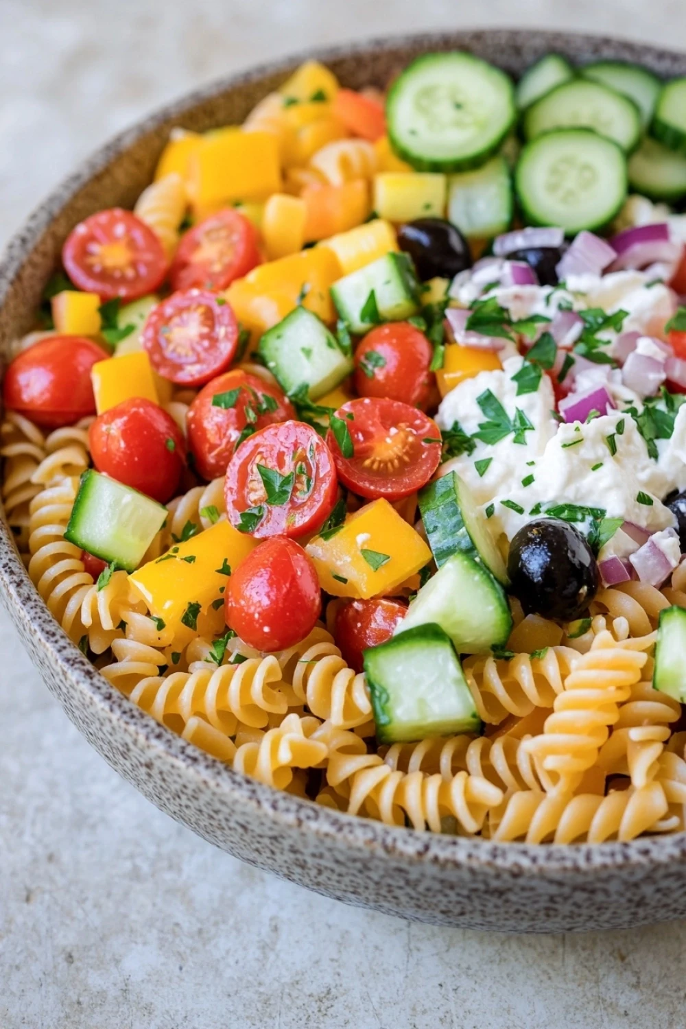 Make ahead pasta salad - the image is a close-up of a bowl of pasta salad. the bowl is made of ceramic and has a light brown color. the pasta is fusilli, which is a type of spiral-shaped pasta, and it is mixed with various vegetables such as sliced cucumbers, red onions, cherry tomatoes, and black olives. the vegetables are arranged in a colorful and appetizing manner, with some of them overlapping each other. there is also a dollop of white dressing on top of the salad, which appears to be feta cheese. the salad is garnished with fresh herbs, likely parsley or cilantro. the background is a white countertop.