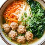 Molly yeh slow cooker asian wedding soup - the image is a close-up of a bowl of soup. the soup appears to be a type of noodle soup with meatballs, carrots, and greens. the meatballs are browned and appear to be seasoned with herbs and spices. the carrots are bright orange and are arranged in a pile on top of the soup. there are also some shredded cheese and chopped green onions scattered throughout the dish. the bowl is made of ceramic and has a brown rim. the background is white.