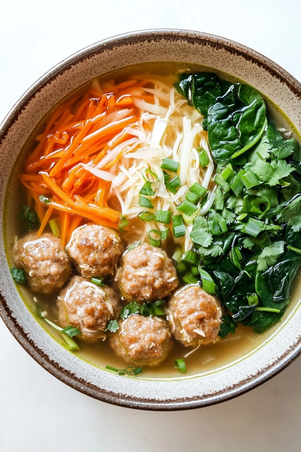 Molly yeh slow cooker asian wedding soup - the image is a close-up of a bowl of soup. the soup appears to be a type of noodle soup with meatballs, carrots, and greens. the meatballs are browned and appear to be seasoned with herbs and spices. the carrots are bright orange and are arranged in a pile on top of the soup. there are also some shredded cheese and chopped green onions scattered throughout the dish. the bowl is made of ceramic and has a brown rim. the background is white.