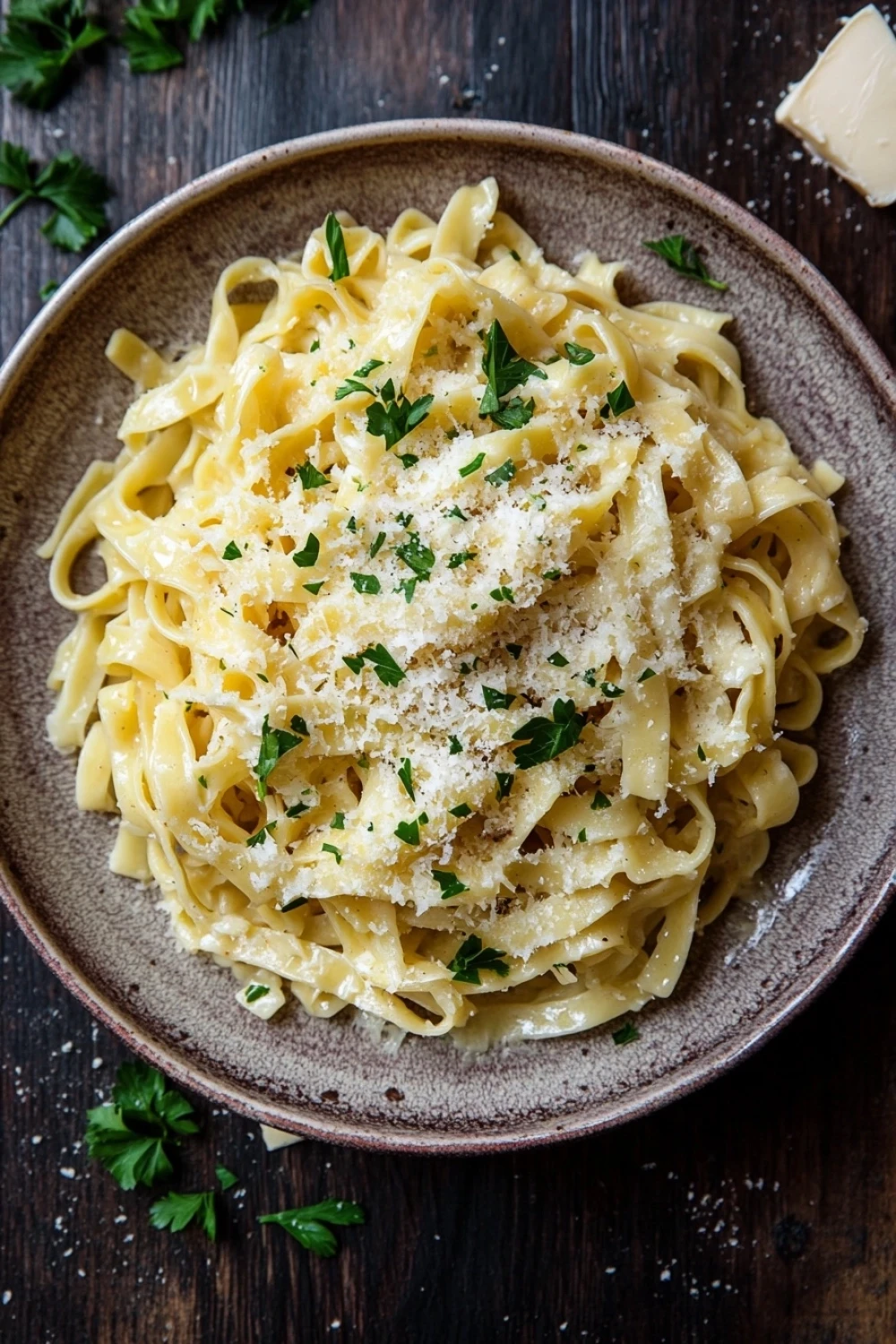 One pot garlic butter noodles - the image shows a plate of fettuccine alfredo pasta on a wooden table. the pasta is cooked al dente and is topped with a generous amount of parmesan cheese and fresh parsley. the plate is brown and has a textured surface. there are a few sprigs of parsley scattered around the plate, adding a pop of green color to the dish. on the right side of the image, there is a small piece of butter, which is partially visible in the top right corner. the background is blurred, but it appears to be a rustic kitchen countertop.
