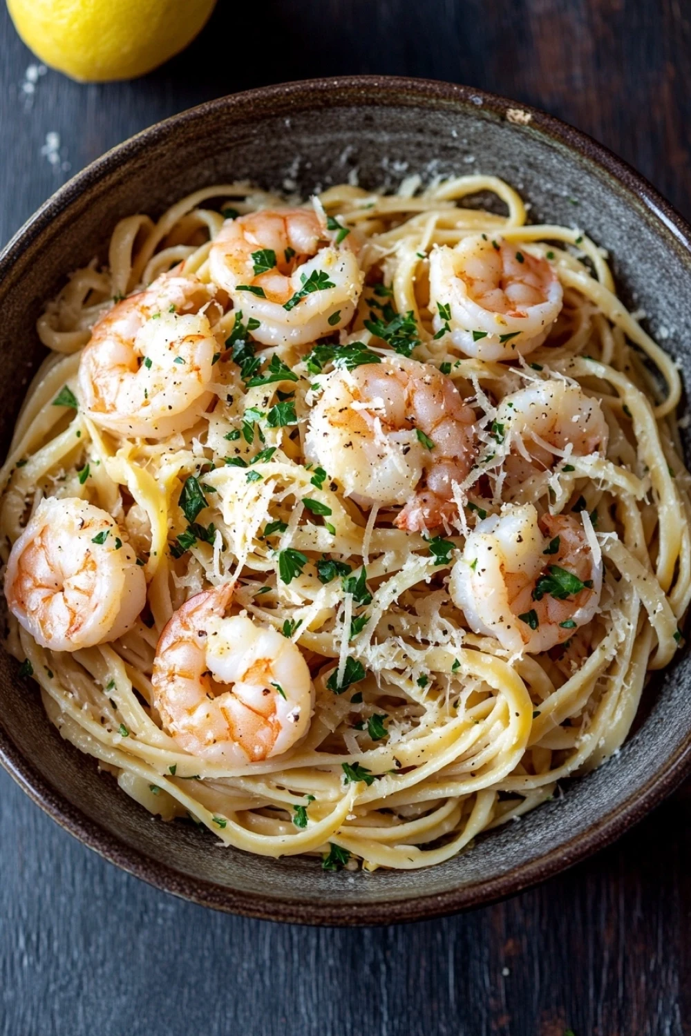 One pot garlic butter shrimp pasta - the image shows a bowl of spaghetti with shrimp and parmesan cheese. the spaghetti is cooked al dente and is garnished with fresh parsley. the shrimp are large and pink, and they are arranged in a circular pattern on top of the spaghetti. the dish is served in a dark brown bowl with a wooden table in the background. there is a lemon wedge on the side of the bowl. the overall color scheme of the dish is warm and inviting.