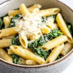 One pot pasta mit spinat - the image is a close-up of a bowl of pasta dish. the pasta appears to be penne, which is a type of penne pasta, and is cooked in a creamy sauce. the dish is garnished with fresh spinach leaves and grated parmesan cheese. the bowl is made of ceramic with a speckled pattern and is sitting on a white marble countertop. the background is blurred, making the pasta and spinach the focal point of the image.