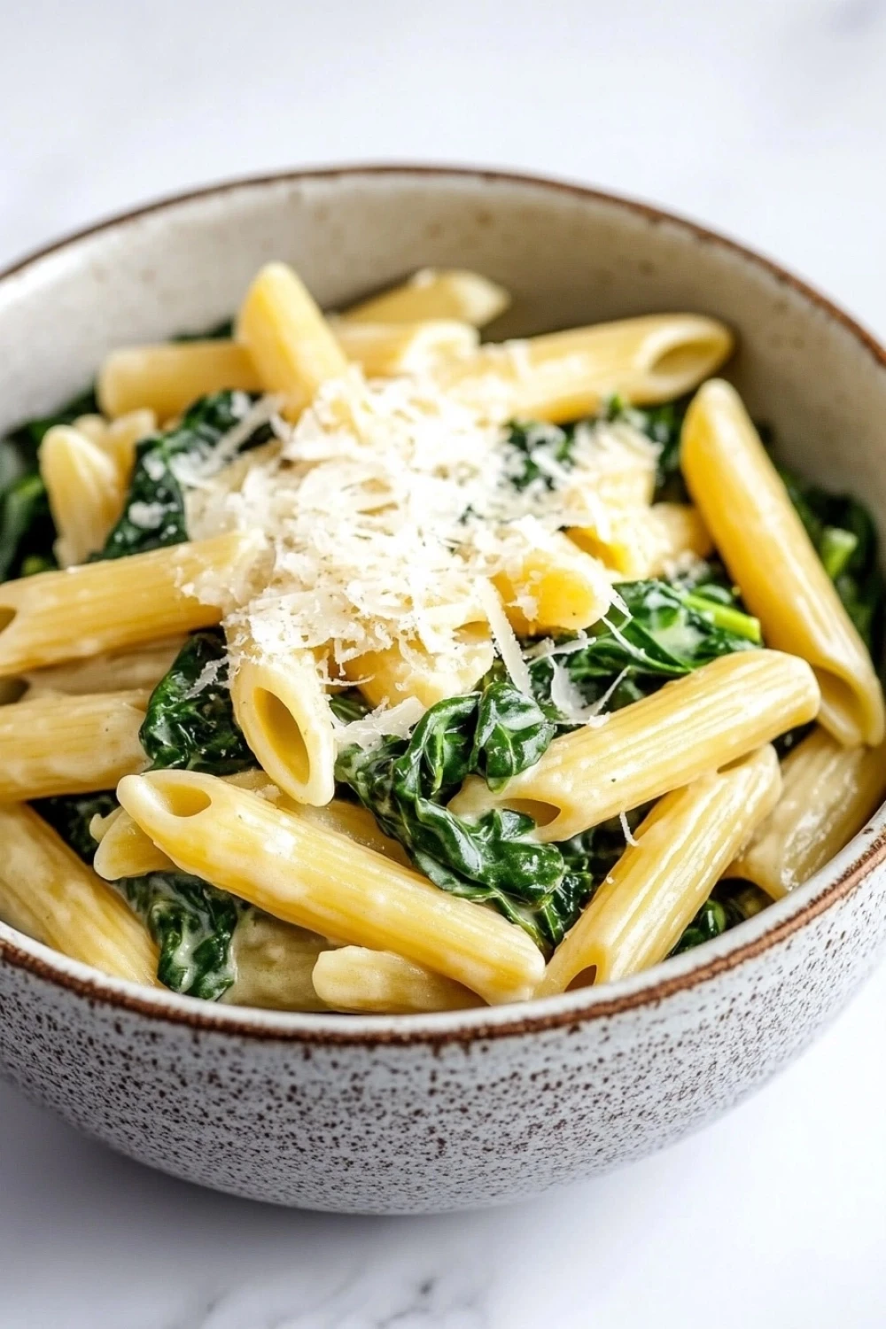 One pot pasta mit spinat - the image is a close-up of a bowl of pasta dish. the pasta appears to be penne, which is a type of penne pasta, and is cooked in a creamy sauce. the dish is garnished with fresh spinach leaves and grated parmesan cheese. the bowl is made of ceramic with a speckled pattern and is sitting on a white marble countertop. the background is blurred, making the pasta and spinach the focal point of the image.