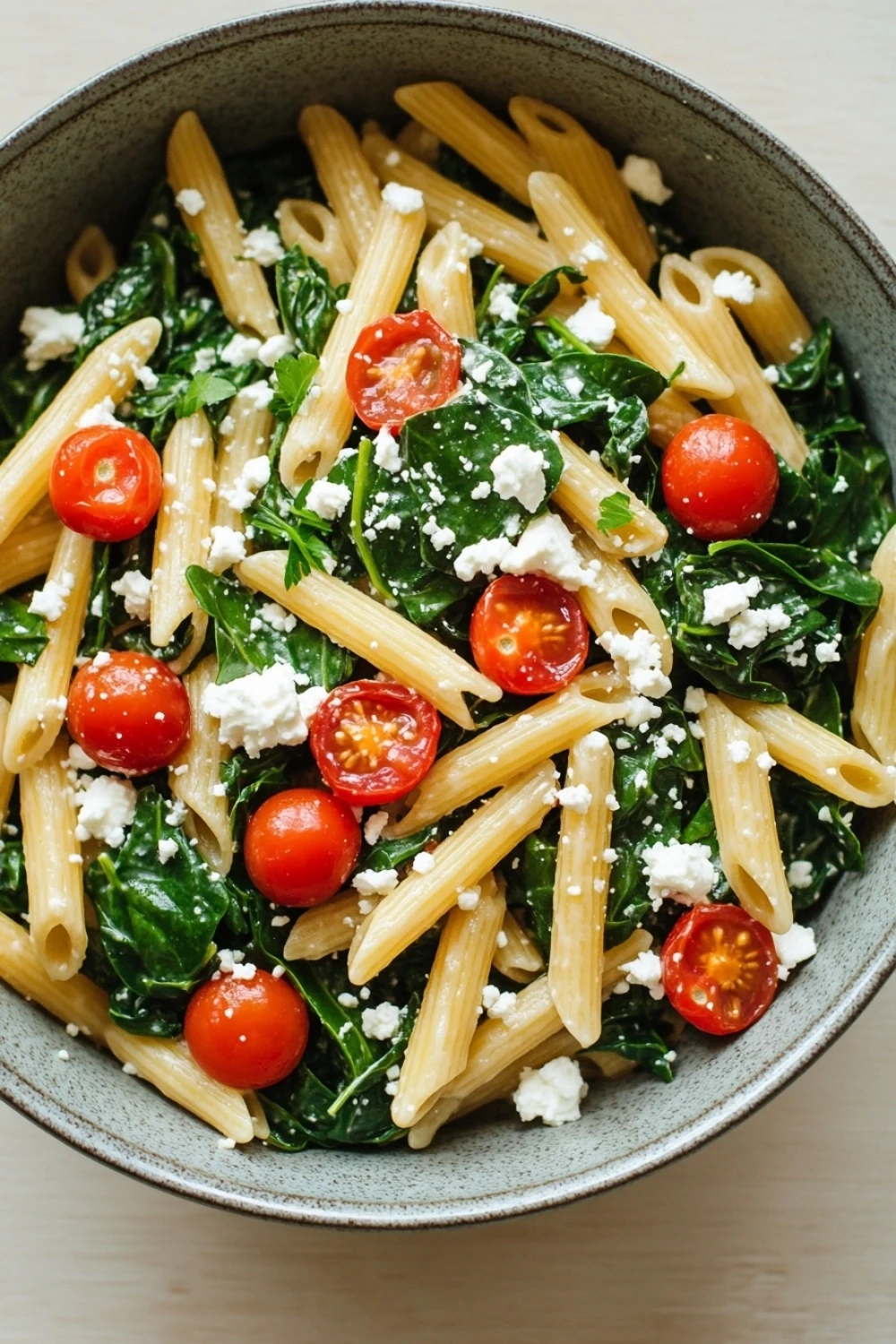 One pot pasta spinat tomaten feta - the image is a close-up of a bowl of pasta salad. the pasta appears to be penne, and it is cooked al dente. the salad is made up of collard greens, cherry tomatoes, and crumbled feta cheese. the tomatoes are bright red and appear to be ripe and juicy. the feta is crumbled and scattered throughout the salad, adding a creamy texture to the dish. the bowl is made of gray ceramic and is sitting on a wooden table.