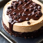 Peanut butter cookie dough cheesecake - the image is of a round cheesecake on a black baking tray. the cheesecake has a creamy white base with a layer of chocolate chips on top. the chocolate chips are scattered across the top of the cheesecake, creating a drizzle of chocolate sauce. the tray is sitting on a dark grey countertop.