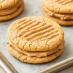 Peanut butter cookie sandwich filling - the image shows a group of freshly baked peanut butter cookies on a baking sheet. the cookies are round and golden brown in color, with a crumbly texture on top. they are arranged in a single layer on the baking sheet, with the top layer being slightly larger than the bottom layer. the peanut butter is spread evenly over the cookies, creating a smooth and creamy texture. a metal spatula is resting on the tray, ready to be used to spread the peanut butter. the background is a white wooden surface.
