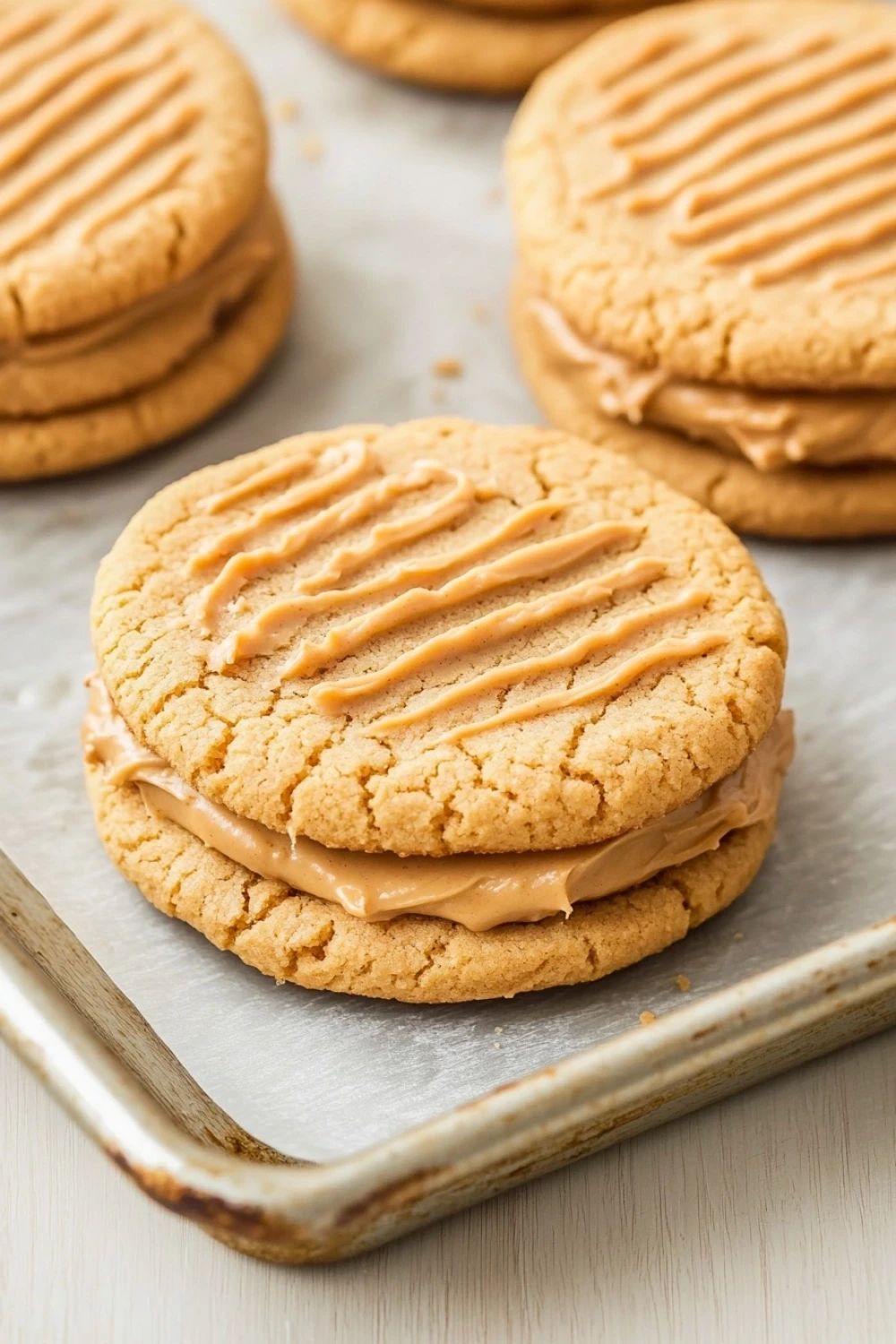 Peanut butter cookie sandwich filling - the image shows a group of freshly baked peanut butter cookies on a baking sheet. the cookies are round and golden brown in color, with a crumbly texture on top. they are arranged in a single layer on the baking sheet, with the top layer being slightly larger than the bottom layer. the peanut butter is spread evenly over the cookies, creating a smooth and creamy texture. a metal spatula is resting on the tray, ready to be used to spread the peanut butter. the background is a white wooden surface.