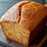 Pumpkin bread high altitude - the image shows a freshly baked loaf of bread on a wooden cutting board. the bread appears to be golden brown in color and has a slightly crumbly texture. it is cut in half, revealing the soft, fluffy interior. the cutting board is placed on a dark grey slate countertop. the background is blurred, making the bread the focal point of the image.
