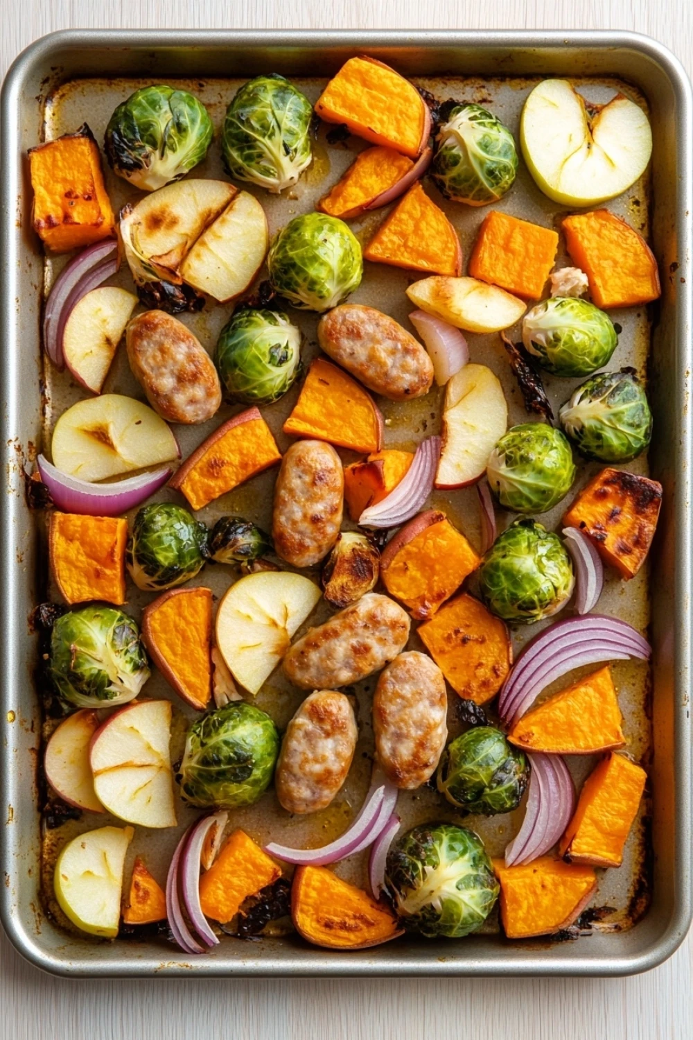 Sheet pan chicken sausage fall - the image shows a baking tray lined with parchment paper. on the tray, there are various types of vegetables, including brussels sprouts, apples, and red onions. the vegetables are arranged in a circular pattern, with some overlapping each other. the apples are cut into small cubes and are placed on top of the vegetables. the red onions are scattered throughout the tray. the tray is sitting on a white wooden surface.
