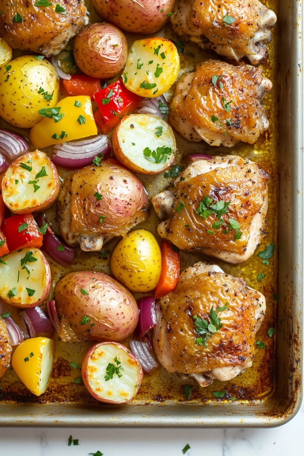 Sheet pan dinner with potatoes - the image shows a baking tray filled with roasted chicken and potatoes. the chicken is golden brown and appears to be seasoned with herbs and spices. the potatoes are cut into small cubes and are arranged in a circular pattern on the tray. there are also diced red and yellow bell peppers, red onions, and yellow potatoes scattered throughout the dish. the tray is lined with parchment paper and there are a few sprigs of parsley on top. the background is a white marble countertop.