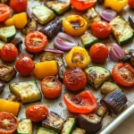 Sheet pan roasted mediterranean vegetables - the image shows a baking tray filled with roasted vegetables. the vegetables are arranged in a circular pattern on the tray, with some overlapping each other. the tray is lined with parchment paper and appears to be on a wooden table. the vegetables include sliced zucchini, cherry tomatoes, bell peppers, and red onions. the tomatoes are bright red and appear to be seasoned with herbs and spices. the bell peppers are yellow and the onions are dark red. the zucchinis are cut into small cubes and are arranged around the tray in a way that they are evenly spaced. the overall appearance of the vegetables is colorful and appetizing.