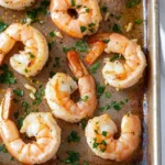 Sheet pan shrimp with garlic - the image shows a baking tray lined with parchment paper. on the tray, there are several cooked shrimp arranged in a single layer. the shrimp are orange in color and appear to be seasoned with herbs and spices. the tray is sitting on a white marble countertop with a white cloth napkin on the side. the background is blurred, but it appears to be a kitchen countertop.