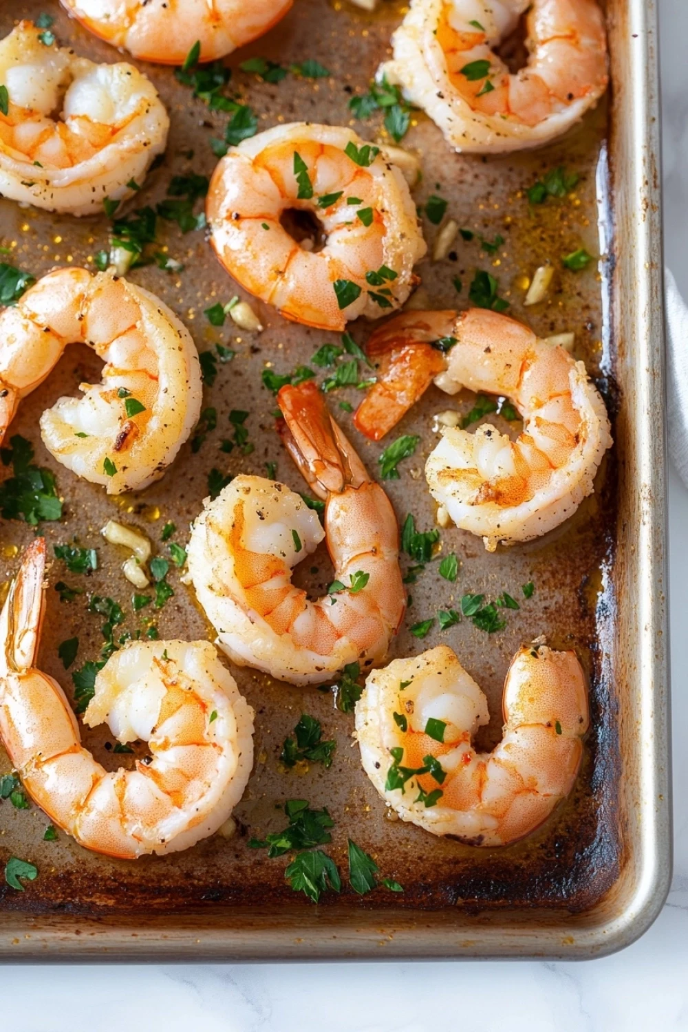 Sheet pan shrimp with garlic - the image shows a baking tray lined with parchment paper. on the tray, there are several cooked shrimp arranged in a single layer. the shrimp are orange in color and appear to be seasoned with herbs and spices. the tray is sitting on a white marble countertop with a white cloth napkin on the side. the background is blurred, but it appears to be a kitchen countertop.